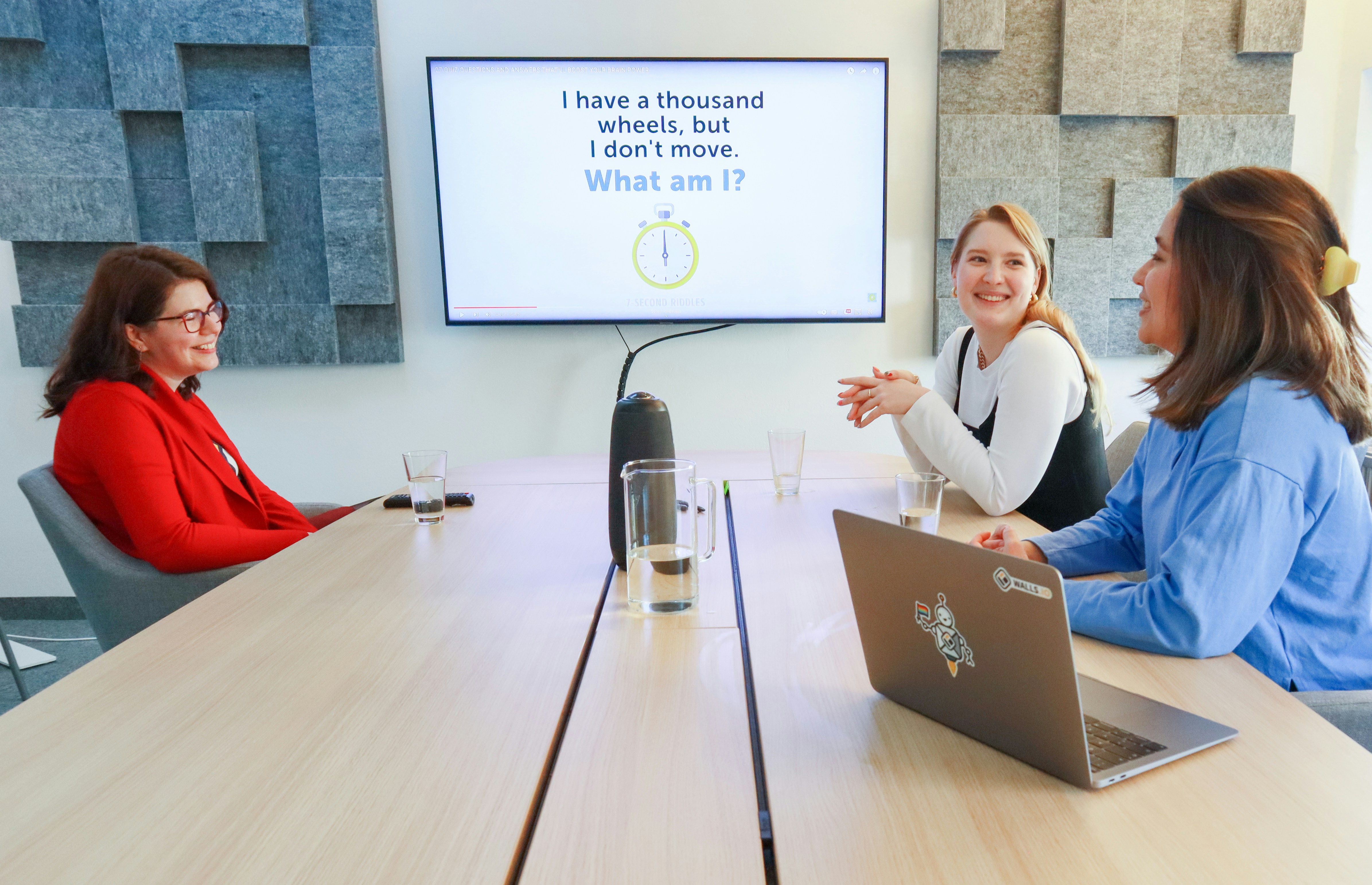 Three colleagues smiling and interacting in a conference room while participating in a quiz displayed on a screen.